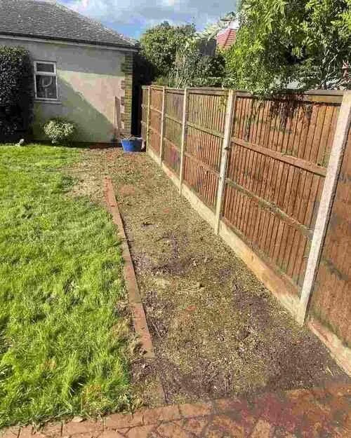 A narrow, cleared strip of soil and debris runs alongside a wooden fence in a garden, with a patch of green grass to the left and a building in the background, illustrating a garden clearance or preparation task often performed by Tree Surgeons in Abridge.