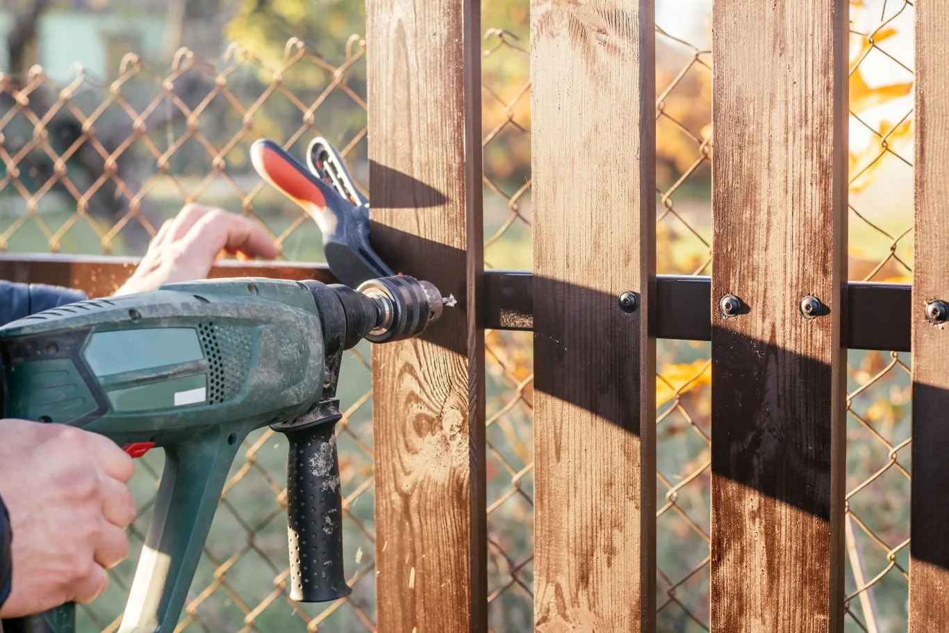 A professional carrying out fence installation services using a power drill to secure wooden fence panels onto a metal frame, with chain link fencing in the background.